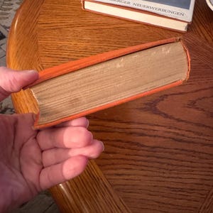 K&ouml;nnte beinhalten: Ein orangefarbenes Buch mit gealterten, hellbraunen Seiten. Das Buch wird in einer Hand gehalten. Ein weiteres Buch mit blauem Einband und dem Text "HEIDELBERGER NEUERWERBUNGEN" ist im Hintergrund zu sehen.
