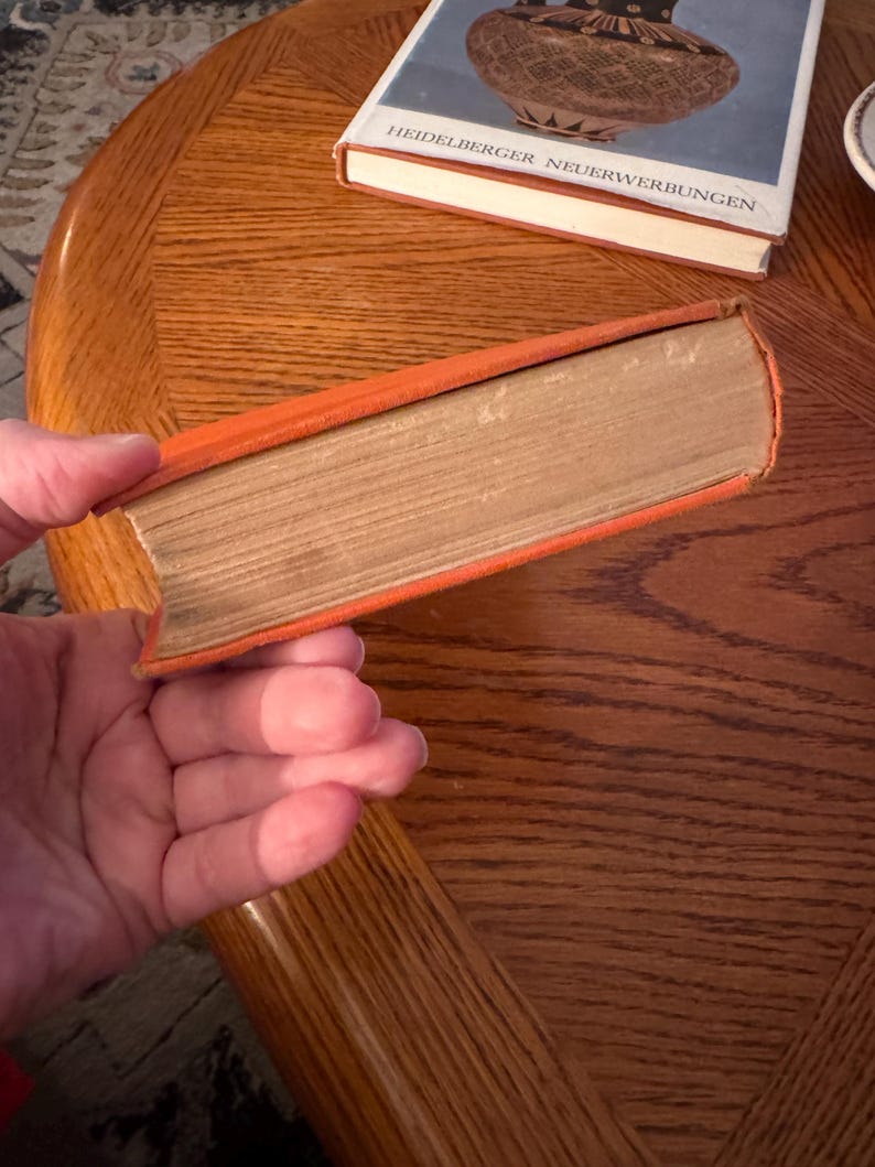 K&ouml;nnte beinhalten: Ein orangefarbenes Buch mit gealterten, hellbraunen Seiten. Das Buch wird in einer Hand gehalten. Ein weiteres Buch mit blauem Einband und dem Text "HEIDELBERGER NEUERWERBUNGEN" ist im Hintergrund zu sehen.