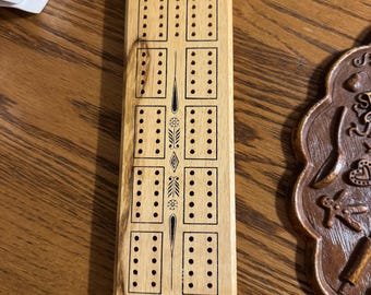 May include: A wooden cribbage board with a light wood grain. The board has multiple rows of holes for pegging, with decorative etched lines and the text "MB A MILTON BRADLEY COMPANY".
