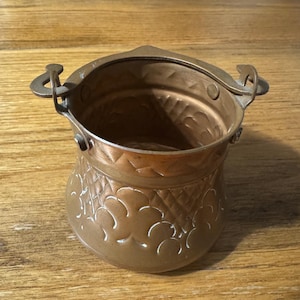 May include: A small, antique copper-colored metal bucket with a handle and decorative floral patterns. The bucket has a rounded base and a flared top, with a handle on each side. The bucket is sitting on a wooden surface.