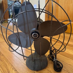 May include: An antique Arctic brand electric fan with four blades. The fan has a dark gray metal cage and base, with a black power cord. The blades are a darker gray color. The fan is sitting on a wooden floor.