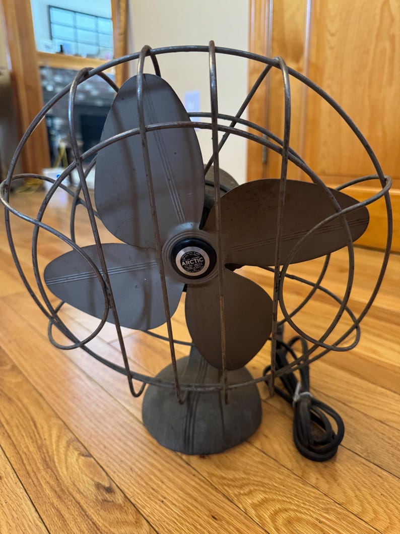 May include: An antique Arctic brand electric fan with four blades. The fan has a dark gray metal cage and base, with a black power cord. The blades are a darker gray color. The fan is sitting on a wooden floor.