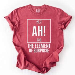 May include: A red t-shirt with rolled-up sleeves. The shirt features a white square with the numbers "29,7" and "230" and the letters "AH!" inside. Below the square, the text reads "THE ELEMENT OF SURPRISE."