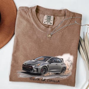 May include: A brown short-sleeve t-shirt featuring a graphic of a grey Toyota GR Corolla car with dust clouds. The shirt has a crew neckline and a Comfort Colors brand label on the inside collar. A silver-toned necklace with a small pendant rests on top.