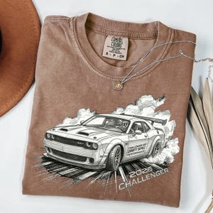 May include: Brown Comfort Colors t-shirt featuring a detailed graphic of a white Dodge Challenger SRT drifting, with the text "2026 Challenger". A silver necklace with a pendant is draped across the shirt.