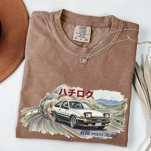 May include: Brown Comfort Colors t-shirt featuring a graphic of a white car drifting on a road with a mountain backdrop. Japanese text is above the car, and the text "AE86 SPRINTER TRUENO" is below. A silver necklace with a pendant is on the shirt.