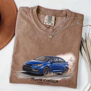 May include: Brown Comfort Colors t-shirt featuring a blue Toyota Corolla sedan graphic. The graphic includes the text "TOYOTA COROLLA SEDAN 2026". A silver necklace with a pendant is draped on the shirt.