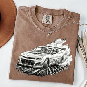 May include: Brown Comfort Colors t-shirt featuring a detailed graphic of a white sports car drifting, with the text "2026 CAMARO" and "CAMARO DRIFT ZL1 PERFORMANCE". A silver necklace with a pendant is draped across the shirt.