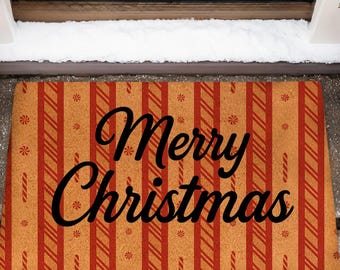 May include: A rectangular doormat with a festive "Merry Christmas" message in black script. The mat features a pattern of vertical red stripes on a tan background. Two small evergreen trees flank the mat, adding to the holiday theme.