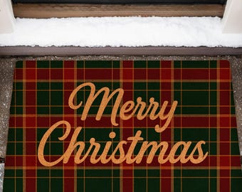 Op de afbeelding: Een rechthoekige deurmat met een rood, groen en goud geruit patroon. De woorden "Merry Christmas" zijn in gouden letters geschreven. De mat ligt voor een zwarte deur, met sneeuw en groenblijvende planten aan weerszijden, wat een feestelijke scène creëert.