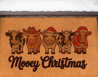 May include: A rectangular doormat with a brown background features five cartoon cows wearing hats and holiday attire. The text "Moovey Christmas" is printed in a dark brown script below the cows. The mat is placed in front of a black door.