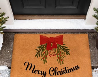 Op de afbeelding: Een rechthoekige deurmat met een feestelijk ontwerp. De mat is bruin met een rode strik en groene takken. De woorden "Merry Christmas" zijn in een sierlijk schrift geschreven. Twee kleine kerstbomen staan aan weerszijden.