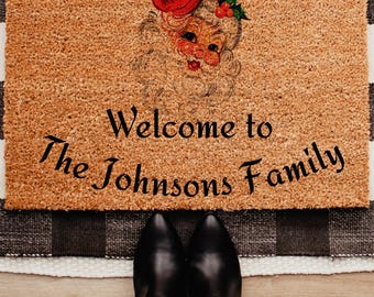 Op de afbeelding: Een rechthoekige deurmat met een illustratie van de kerstman en de tekst "Welcome to The Johnsons Family". De mat is bruin met een zwart-wit geblokte rand. Zwarte laarzen zijn zichtbaar aan de onderkant van de afbeelding.