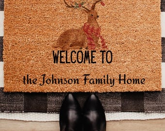 Op de afbeelding: Een rechthoekige deurmat met een hertenillustratie met een sjaal en geweien met bessen. De tekst "WELCOME TO the Johnson Family Home" is in het zwart gedrukt. De mat is gemaakt van een bruin, getextureerd materiaal.