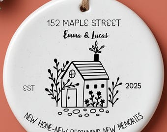 May include: A white ceramic ornament with a house illustration and the text "152 Maple Street, Emma & Lucas, EST 2025, New Home - New Beginning - New Memories." The ornament hangs from a twine string, with a sprig of frosted greenery in the background.
