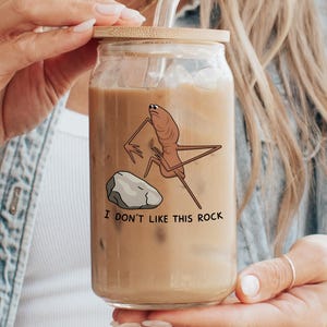 May include: A clear glass tumbler with a bamboo lid and a glass straw. The tumbler features a cartoon illustration of a creature and a rock with the text "I DON'T LIKE THIS ROCK". The tumbler is filled with a brown liquid.