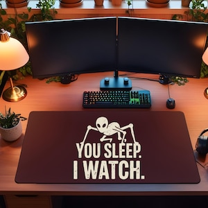May include: A computer desk setup with two monitors, a keyboard, and a mouse. A maroon mousepad with the text "YOU SLEEP. I WATCH." is centered on the desk. Two gold lamps flank the monitors. Headphones are on the right.