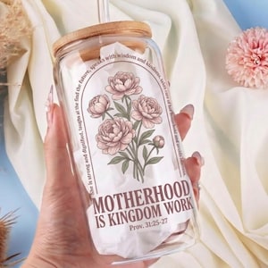 May include: A clear glass tumbler with a wooden lid and a glass straw. The tumbler features a floral design with the text "MOTHERHOOD IS KINGDOM WORK" and a Bible verse. The tumbler is held in a hand.