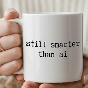 Op de afbeelding: Witte keramische mok met de tekst "still smarter than ai" in het zwart. De mok wordt vastgehouden door een persoon die een witte trui draagt. De mok heeft een wit handvat en een zilveren ring.