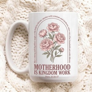 Könnte beinhalten: Weißer Keramikbecher mit großem Henkel. Der Becher zeigt ein Blumendesign aus rosa Pfingstrosen und den Text "MOTHERHOOD IS KINGDOM WORK" mit einem Bibelvers-Verweis. Der Becher steht vor einem weißen, strukturierten Hintergrund.