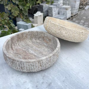 Op de afbeelding: Twee stenen kommen met getextureerde buitenkanten en verschillende interieurpatronen. Eén is rond, de andere ovaal. De kommen zijn lichtbeige en grijs en staan op een marmeren oppervlak. De achtergrond bevat stenen blokken en groen.