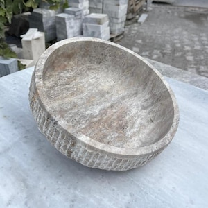 May include: A round, stone bowl with a textured exterior and a smooth interior. The bowl is a light beige color with darker brown and gray veining. The bowl is sitting on a white marble surface.