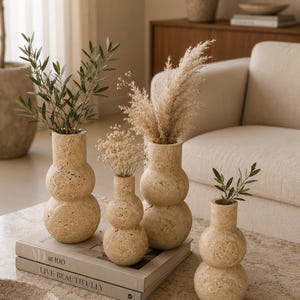 Puede incluir: Cuatro jarrones de travertino beige con una silueta apilada de doble esfera. Los jarrones presentan una textura de piedra natural porosa y contienen varios tallos bot&aacute;nicos secos. Est&aacute;n dispuestos sobre un libro titulado AD at 100 LIVE BEAUTIFULLY.