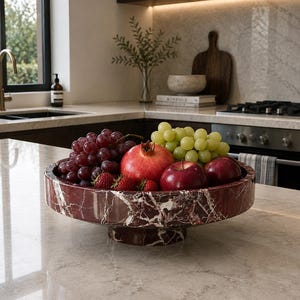 May include: A round, pedestal-style fruit bowl crafted from deep red marble with prominent white veining. The bowl is filled with a variety of fresh produce, including dark purple grapes, green grapes, a bright red pomegranate, and red apples.