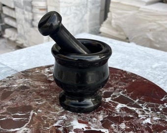 Puede incluir: Un juego de mortero y maja de granito negro. El mortero es redondo con un borde acampanado y una base de pedestal. La maja es cilíndrica con un extremo redondeado. El conjunto está sobre una superficie de mármol rojo y blanco.