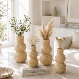 Puede incluir: Cuatro jarrones de piedra travertino beige con una silueta de doble esfera apilada. Los jarrones contienen varios elementos bot&aacute;nicos secos, incluyendo hojas verdes, paniculata blanca, hierba de la pampa y c&aacute;psulas de amapola. Hay dos libros titulados LIVE BEAUTIFULLY.