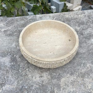 K&ouml;nnte beinhalten: Eine runde, beige Steinschale mit einer strukturierten, geriffelten Au&szlig;enseite. Die Schale hat einen glatten Innenraum und steht auf einer grauen Oberfl&auml;che. Die Schale dient wahrscheinlich dekorativen Zwecken oder als Servierschale.