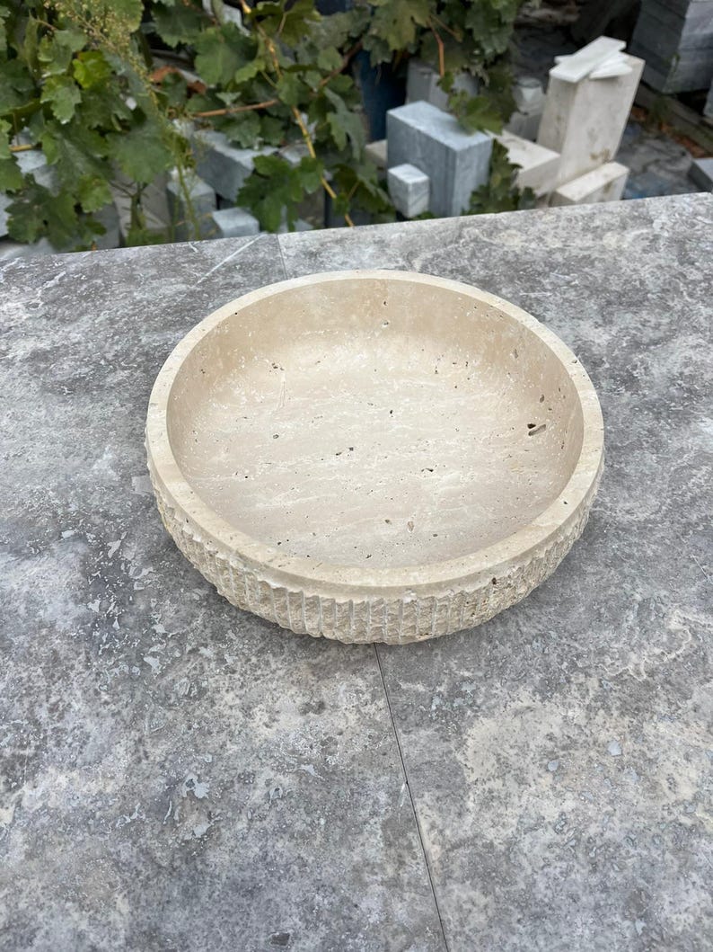 K&ouml;nnte beinhalten: Eine runde, beige Steinschale mit einer strukturierten, geriffelten Au&szlig;enseite. Die Schale hat einen glatten Innenraum und steht auf einer grauen Oberfl&auml;che. Die Schale dient wahrscheinlich dekorativen Zwecken oder als Servierschale.