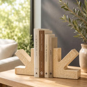 May include: Two beige, textured stone bookends with an angular, arrow-like shape holding four books titled The Kinfolk Home, Arranging Things, Live Beautifully, and at 100. The bookends have a porous, natural finish and are positioned on a wooden surface.
