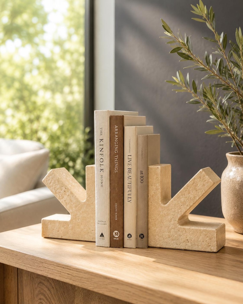 May include: Two beige, textured stone bookends with an angular, arrow-like shape holding four books titled The Kinfolk Home, Arranging Things, Live Beautifully, and at 100. The bookends have a porous, natural finish and are positioned on a wooden surface.