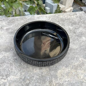 May include: A round, black bowl with a textured rim. The bowl has a glossy interior with a white vein-like pattern. The bowl is on a grey surface.