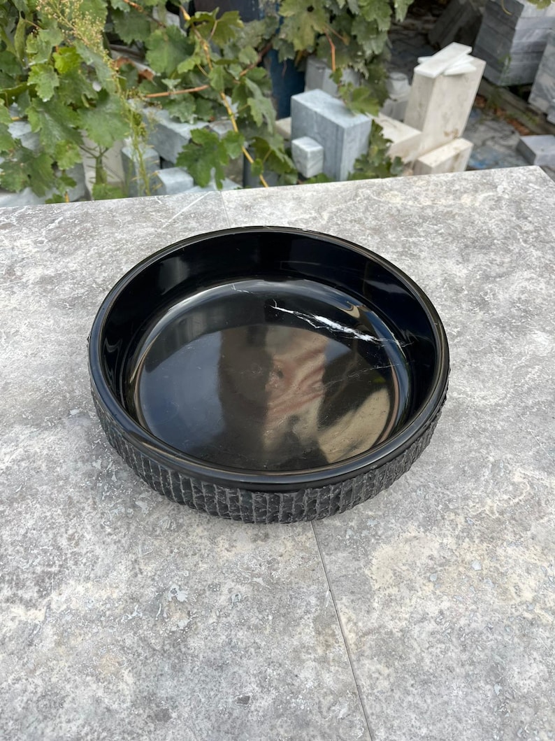 May include: A round, black bowl with a textured rim. The bowl has a glossy interior with a white vein-like pattern. The bowl is on a grey surface.