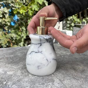 Puede incluir: Un dispensador de jab&oacute;n de m&aacute;rmol con una bomba dorada. El dispensador es blanco con vetas negras. La bomba dispensa l&iacute;quido sobre una mano. El dispensador est&aacute; sobre una superficie gris.