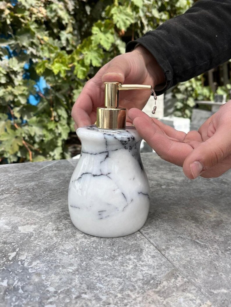 Puede incluir: Un dispensador de jab&oacute;n de m&aacute;rmol con una bomba dorada. El dispensador es blanco con vetas negras. La bomba dispensa l&iacute;quido sobre una mano. El dispensador est&aacute; sobre una superficie gris.