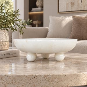 May include: A shallow, circular white stone bowl with a smooth, polished finish and subtle beige veining. The bowl is elevated by three spherical ball-shaped feet made of the same white stone material, providing a stable base for the vessel.