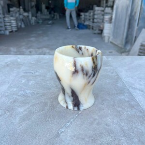 May include: A small, rounded cup carved from polished stone, featuring a smooth, cream-coloured base with prominent, swirling dark brown and black veins throughout the material. The vessel has a wide, open top and a slightly tapered, flared base.