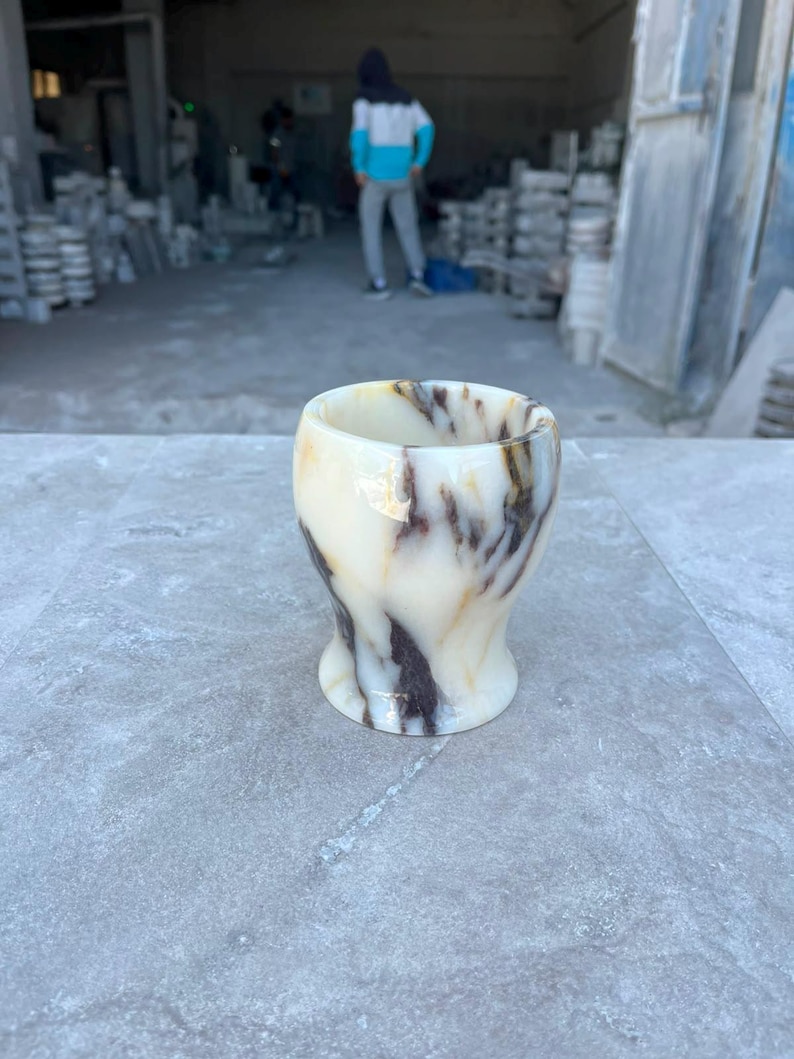 May include: A small, rounded cup carved from polished stone, featuring a smooth, cream-coloured base with prominent, swirling dark brown and black veins throughout the material. The vessel has a wide, open top and a slightly tapered, flared base.