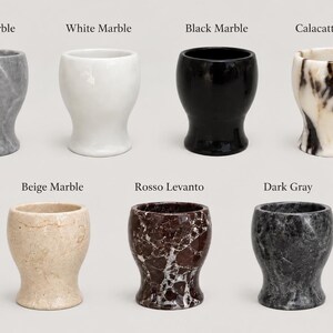 May include: Seven stone egg cups arranged in two rows, labelled Gray Marble, White Marble, Black Marble, Calacatta Viola, Beige Marble, Rosso Levanto, and Dark Gray. Each cup features a smooth, rounded, pedestal-style base and a wide opening.