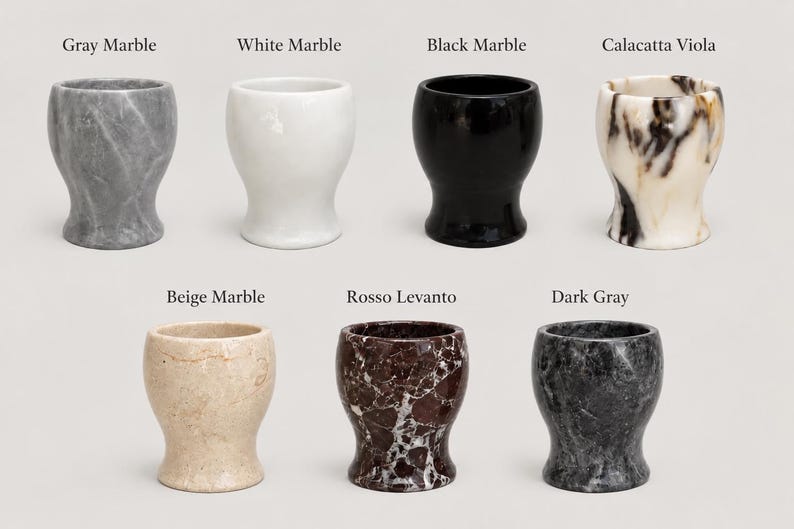 May include: Seven stone egg cups arranged in two rows, labelled Gray Marble, White Marble, Black Marble, Calacatta Viola, Beige Marble, Rosso Levanto, and Dark Gray. Each cup features a smooth, rounded, pedestal-style base and a wide opening.