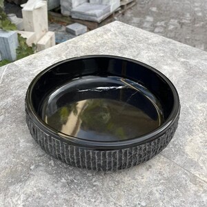May include: A black, round bowl with a textured, vertical-line pattern around the exterior. The interior of the bowl is smooth and reflective. The bowl is on a grey surface.