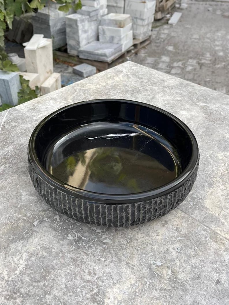 May include: A black, round bowl with a textured, vertical-line pattern around the exterior. The interior of the bowl is smooth and reflective. The bowl is on a grey surface.
