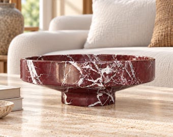 May include: A deep red marble bowl with prominent white veining throughout. The bowl features a wide, shallow basin and a sturdy, circular pedestal base, showcasing the natural polished stone texture and intricate patterns of the dark red material.