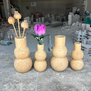 Puede incluir: Cuatro jarrones de travertino beige con una silueta apilada de doble esfera. Dos jarrones contienen c&aacute;psulas de amapola secas y una rosa artificial rosa con hojas verdes. La piedra tiene una superficie porosa y texturizada con variaciones.