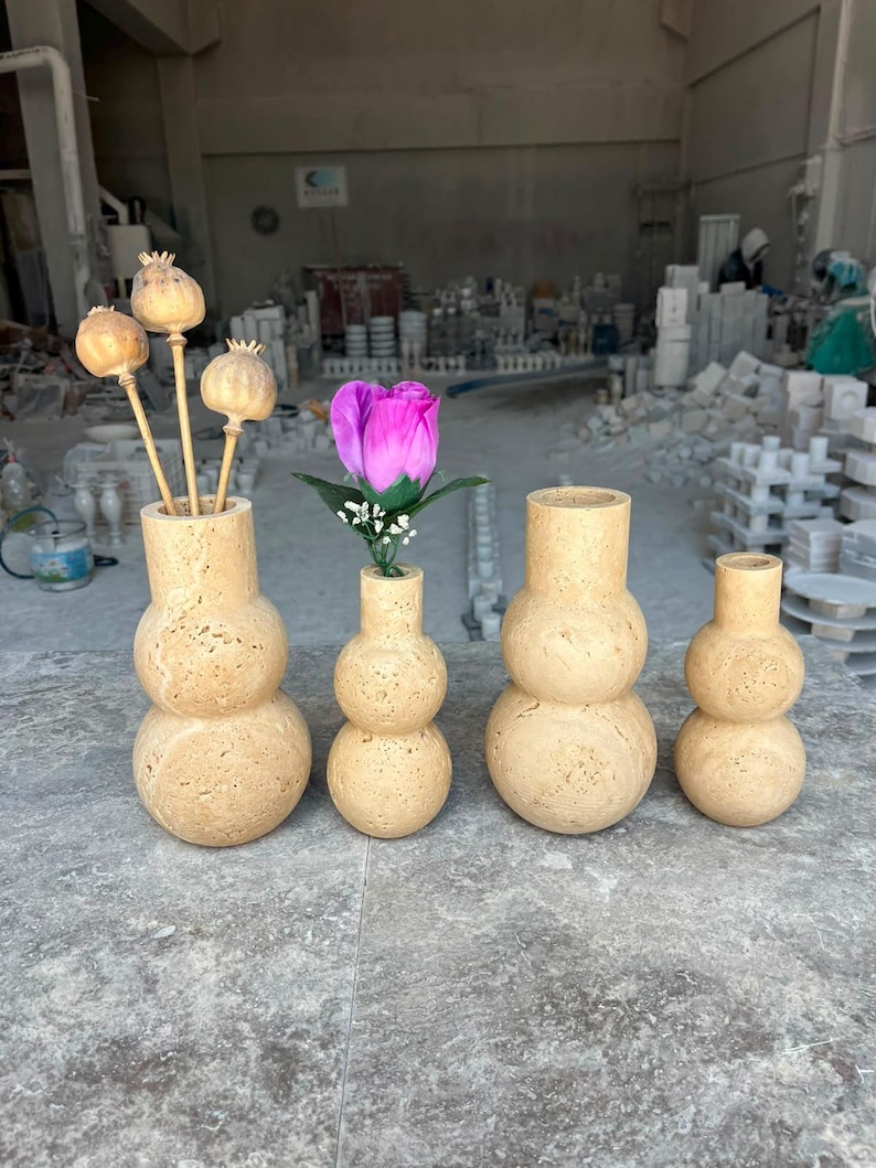 Puede incluir: Cuatro jarrones de travertino beige con una silueta apilada de doble esfera. Dos jarrones contienen c&aacute;psulas de amapola secas y una rosa artificial rosa con hojas verdes. La piedra tiene una superficie porosa y texturizada con variaciones.