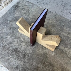 May include: A pair of beige, porous stone bookends with a Y-shaped design, supporting a single dark brown hardcover book. The bookends feature a textured, natural surface with visible small pits and crevices, providing a sturdy base for the book.