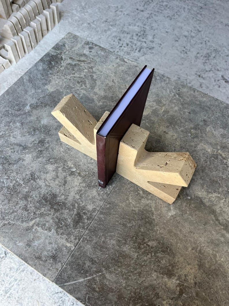May include: A pair of beige, porous stone bookends with a Y-shaped design, supporting a single dark brown hardcover book. The bookends feature a textured, natural surface with visible small pits and crevices, providing a sturdy base for the book.